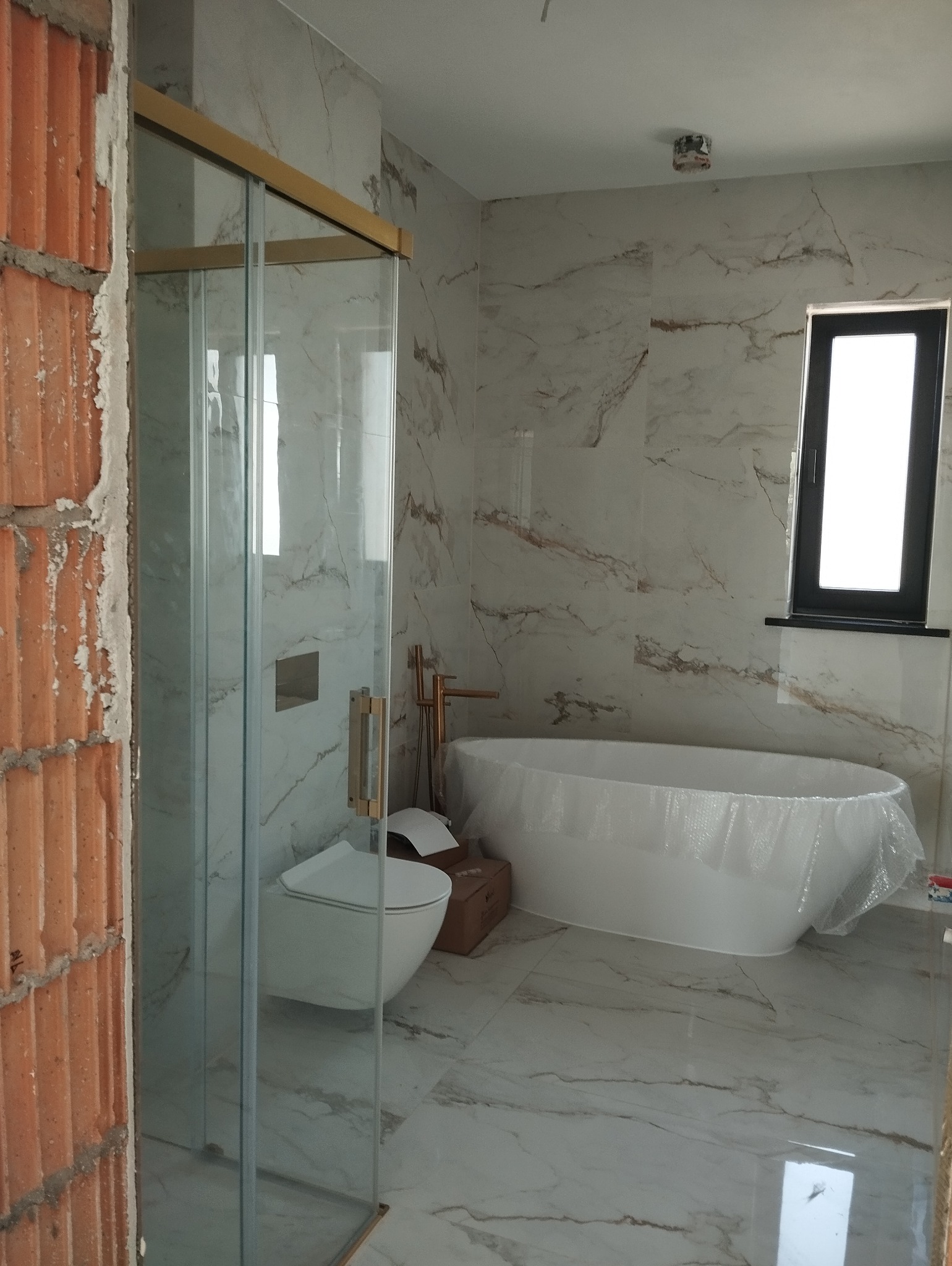 Niedokończona łazienka z marmurowymi płytkami, widoczna kabina prysznicowa ze złotymi ramami, podwieszana toaleta, wolnostojąca wanna owinięta folią ochronną i fragment ściany z surowej cegły.