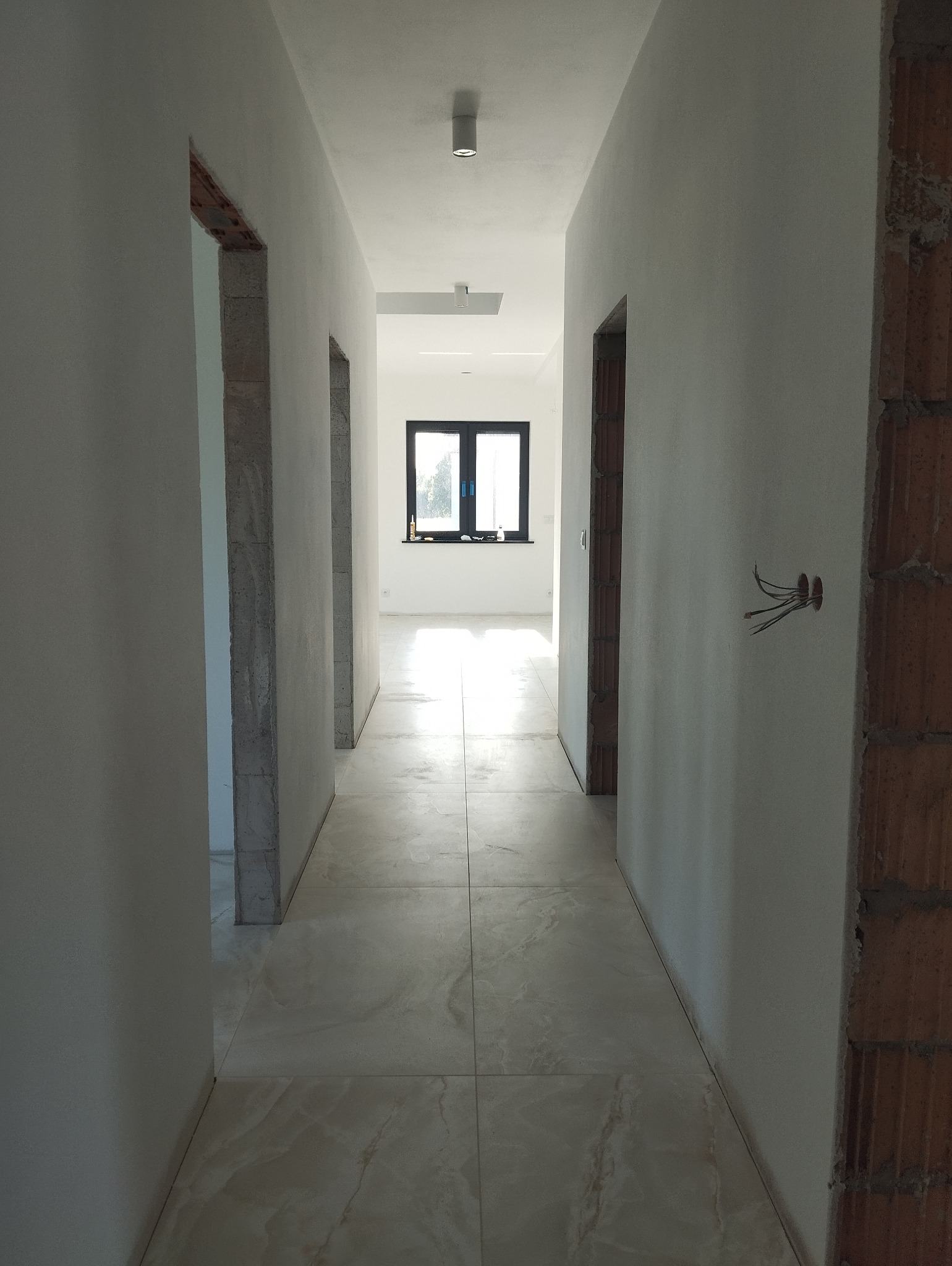 Długi korytarz w stanie surowym z położoną jasną podłogą z płytek imitujących marmur, widoczne przygotowane otwory drzwiowe, ściany po gładziach, instalacja elektryczna wystająca ze ściany, okno...