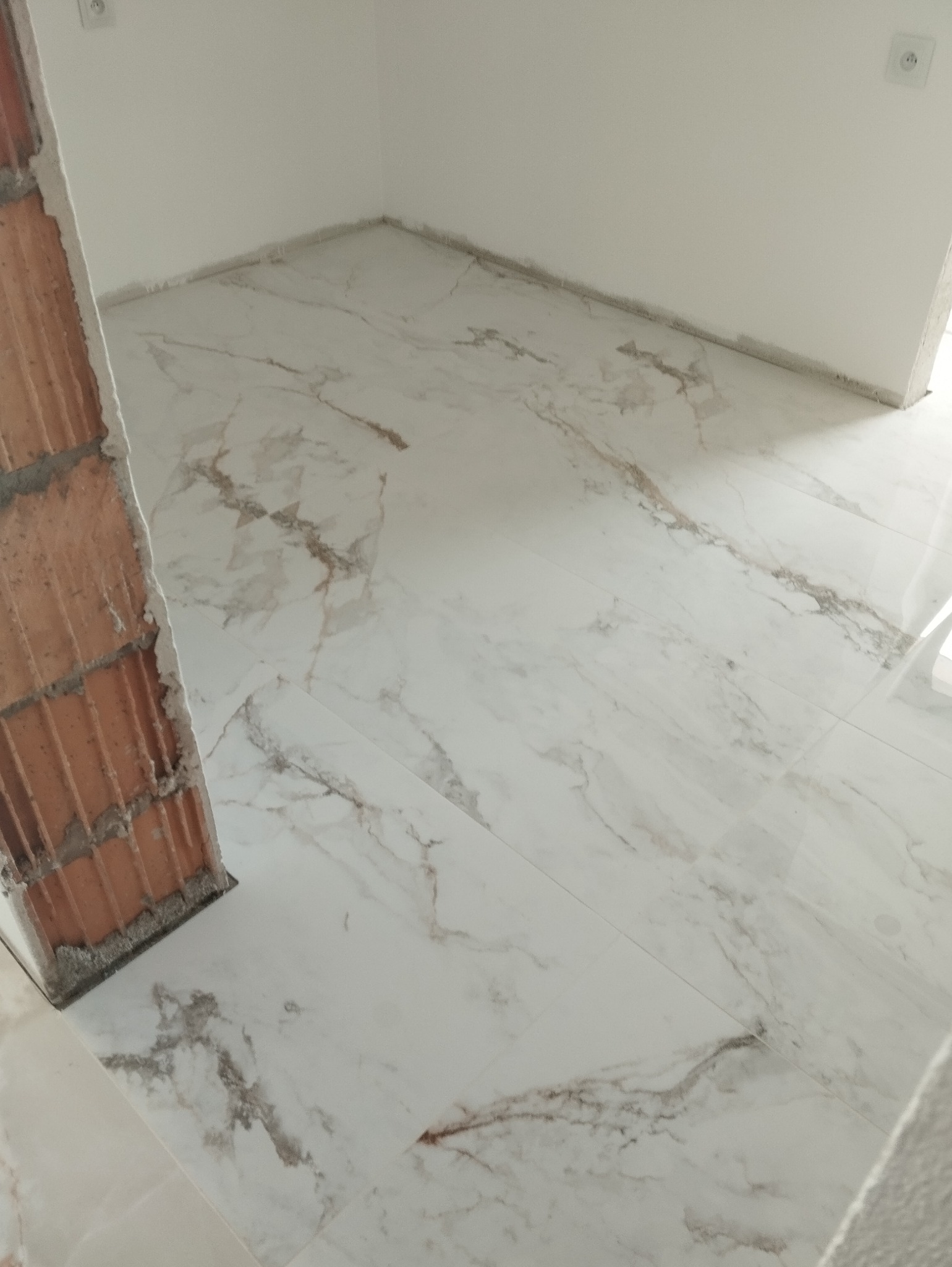 Układanie białych płytek imitujących marmur w łazience, widoczny fragment surowego muru z cegieł i gniazdko elektryczne na ścianie.