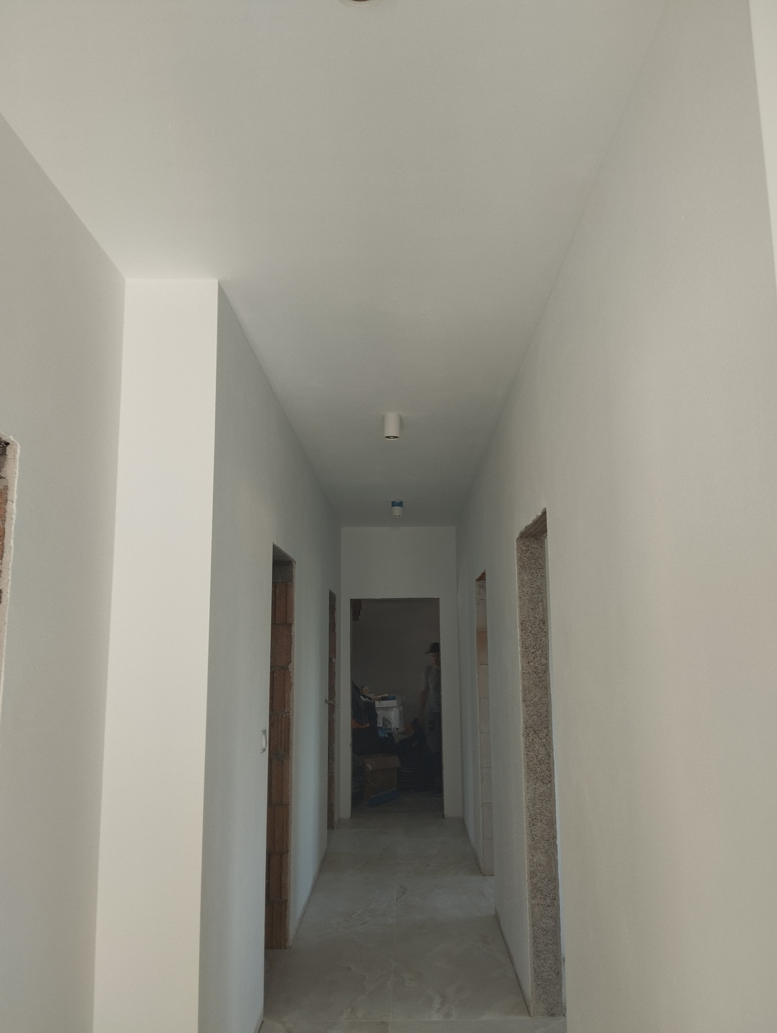 Korytarz w trakcie odświeżania, ściany pomalowane na biało, widoczne surowe obramowania drzwi i fragmenty instalacji oświetleniowej na suficie.