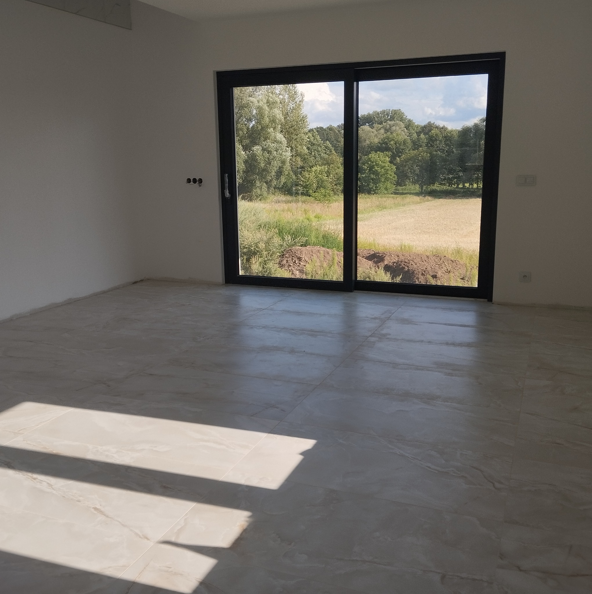 Wnętrze pustego pokoju z nową, jasną, marmurkową podłogą i dużym przesuwnym oknem balkonowym z widokiem na zewnątrz, gdzie widać trawę i drzewa.