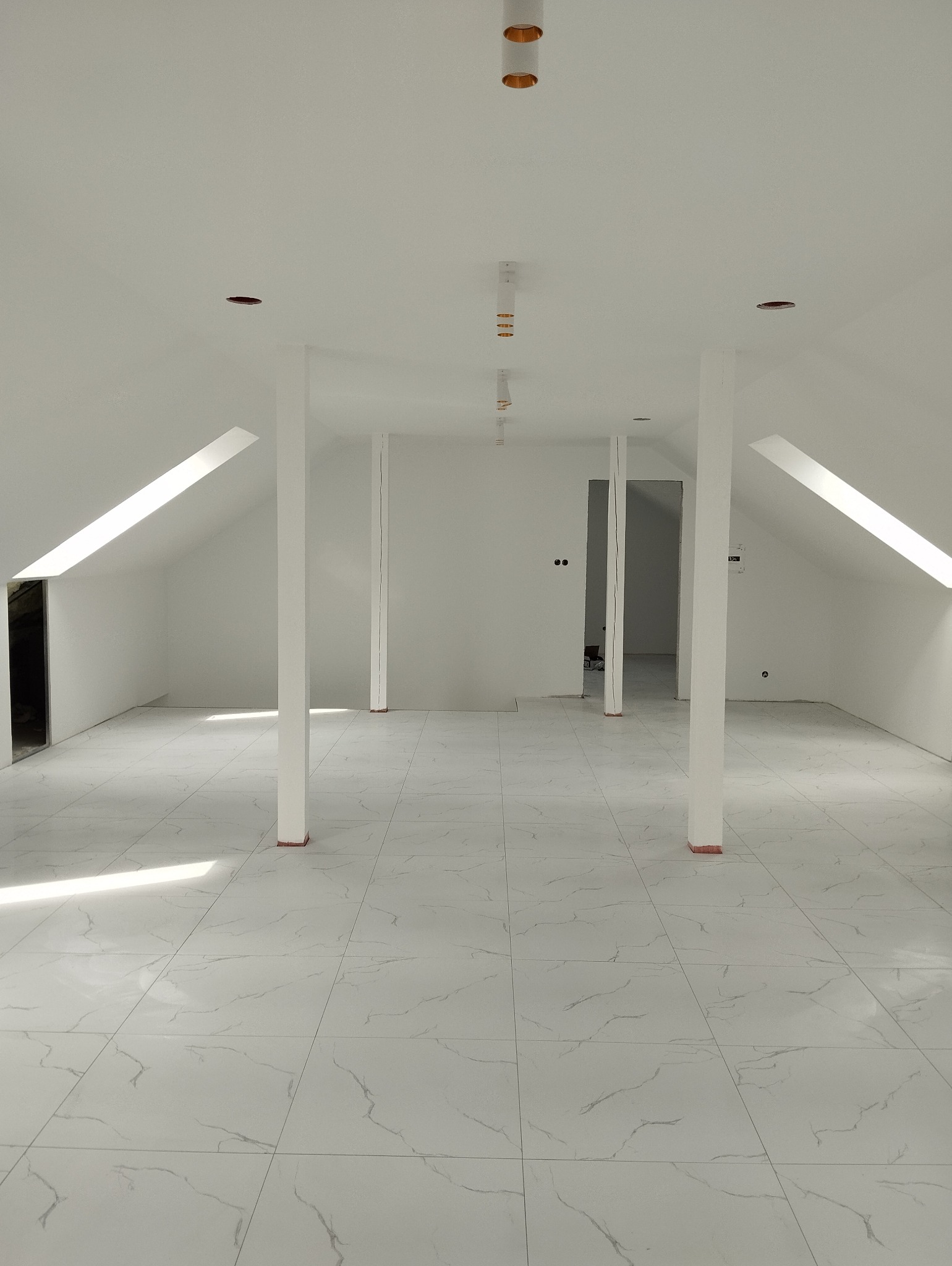 Przestronne, jasne poddasze z białymi ścianami, marmurową posadzką i filarami, przygotowane do dalszych prac wykończeniowych, widoczne otwory na oświetlenie sufitowe.