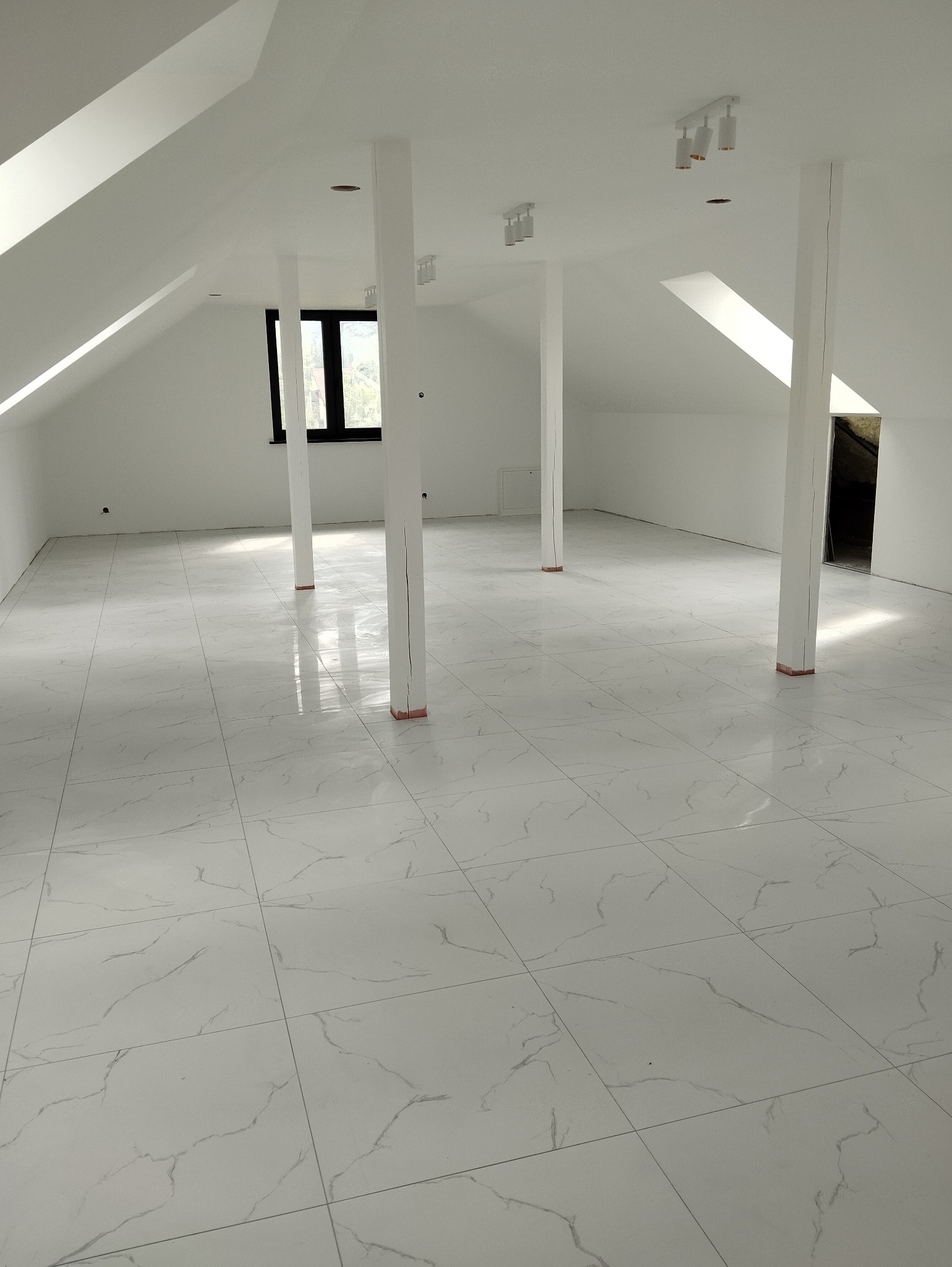 Poddasze po remoncie z białymi ścianami, kolumnami i marmurową podłogą z płytek, widoczne czarne ramy okienne.