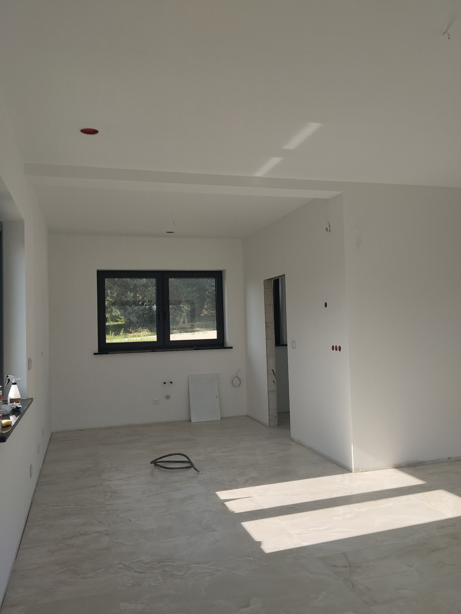 Jasne wnętrze w trakcie wykończenia: świeżo pomalowane białe ściany, okno z czarną ramą, marmurowe płytki podłogowe, widoczne instalacje elektryczne i otwór drzwiowy.