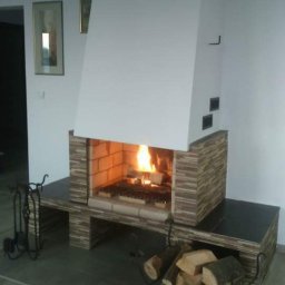 PIMMAR SPÓŁKA Z OGRANICZONĄ ODPOWIEDZIALNOŚCIĄ - Nowoczesny kominek z wkładem obudowanym płytkami imitującymi kamień, z paleniskiem wypełnionym płonącym drewnem, na podłodze stojak z drewnem, a na ścianie obrazy w złotych ramach.