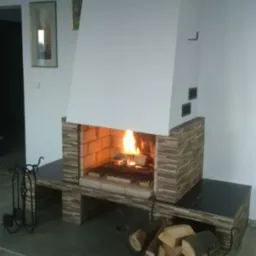 Nowoczesny kominek z wkładem obudowanym płytkami imitującymi kamień, z paleniskiem wypełnionym płonącym drewnem, na podłodze stojak z drewnem, a na ścianie obrazy w złotych ramach.