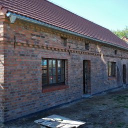 PIMMAR SPÓŁKA Z OGRANICZONĄ ODPOWIEDZIALNOŚCIĄ - Surowa, czerwona elewacja z cegły klinkierowej nowo wybudowanego domu z widocznymi oknami i drzwiami, częściowo zasłonięta rusztowaniem, z fragmentem dachu z czerwonej dachówki i widoczną rynną.