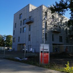 PIMMAR SPÓŁKA Z OGRANICZONĄ ODPOWIEDZIALNOŚCIĄ - Wielopiętrowy budynek mieszkalny z szarej elewacji panelowej, z małymi balkonami, widoczny zza ogrodzenia budowy i kontenera z logo firmy.