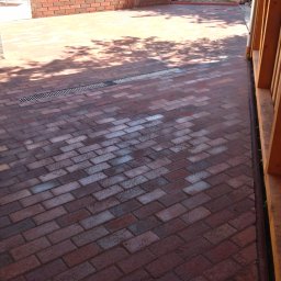 PIMMAR SPÓŁKA Z OGRANICZONĄ ODPOWIEDZIALNOŚCIĄ - Murarz w niebieskiej koszulce w trakcie wznoszenia niskiego muru z czerwonej cegły na zewnątrz budynku, widok z góry na brukowaną powierzchnię.