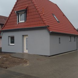 PIMMAR SPÓŁKA Z OGRANICZONĄ ODPOWIEDZIALNOŚCIĄ - Nowoczesny dom z szarą elewacją i czerwoną dachówką, widoczne okna dachowe i rynny, teren wokół domu częściowo wyłożony kostką brukową, a częściowo przygotowany pod trawnik.