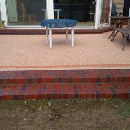 PIMMAR SPÓŁKA Z OGRANICZONĄ ODPOWIEDZIALNOŚCIĄ - Wykończony taras z żywicy epoksydowej w kolorze brzoskwini, z dwustopniowymi schodami z czerwonej cegły, białymi drzwiami tarasowymi i meblami ogrodowymi.