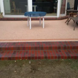 Wykończony taras z żywicy epoksydowej w kolorze brzoskwini, z dwustopniowymi schodami z czerwonej cegły, białymi drzwiami tarasowymi i meblami ogrodowymi.