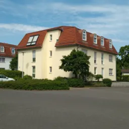 Żółty budynek mieszkalny z czerwonym dachem, widoczne okna dachowe i lukarny, otoczony zielenią, na pierwszym planie brukowany plac.