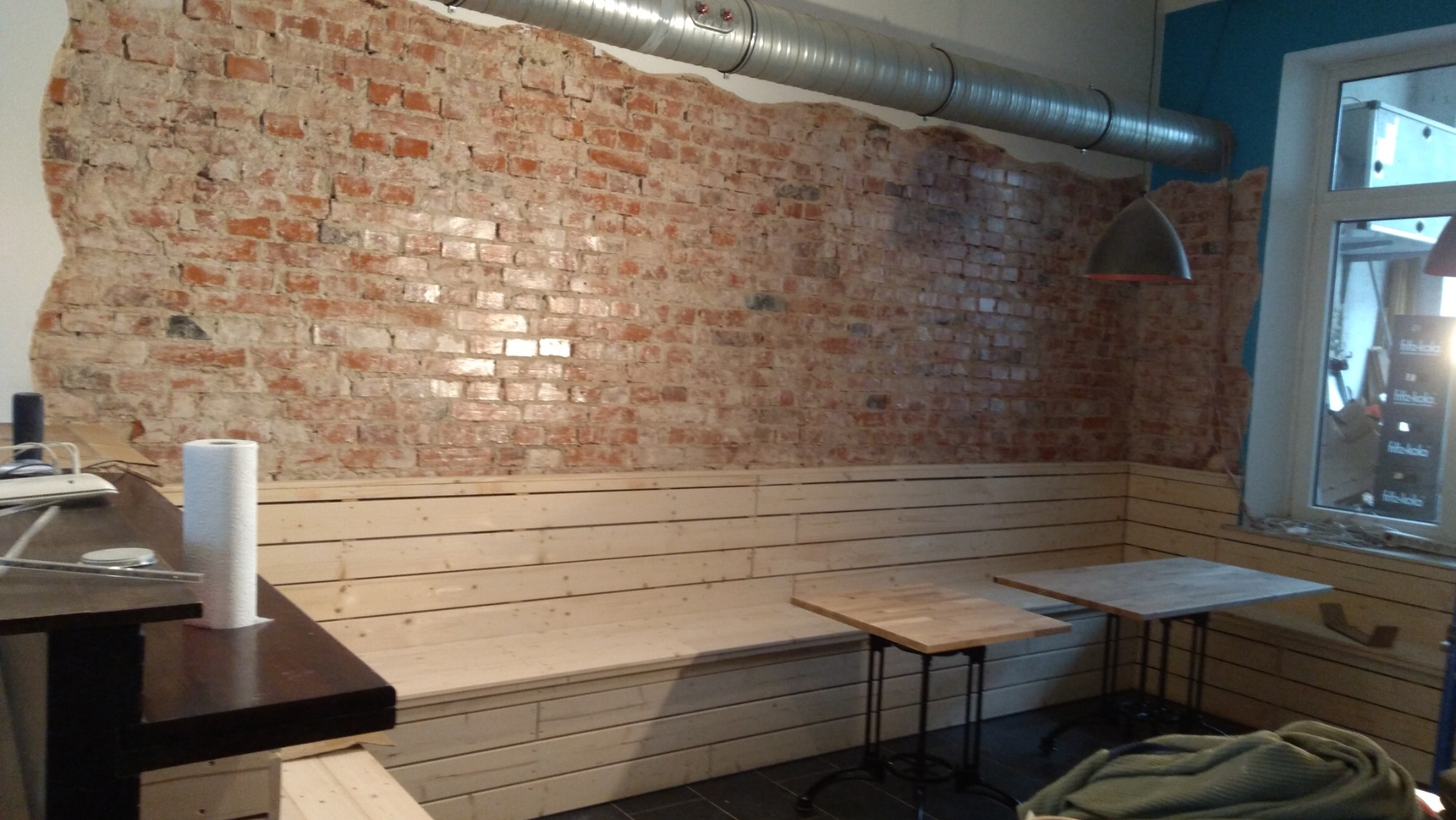 Surowa, ceglana ściana z fragmentami starego tynku, połączona z drewnianą boazerią i ławką, uzupełniona o metalowy kanał wentylacyjny pod sufitem i proste stoliki w kawiarni.