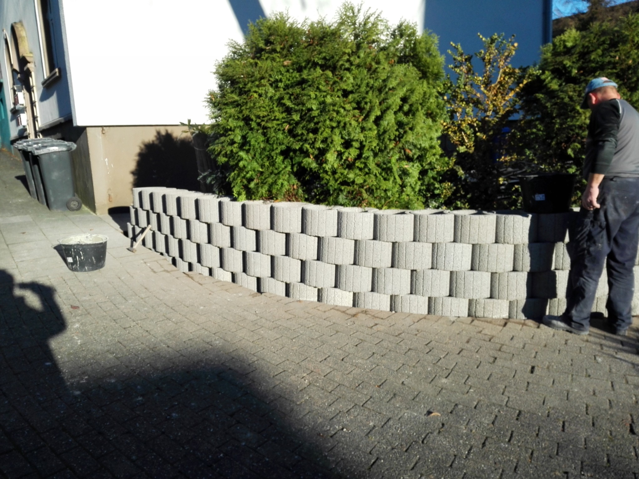 Wznoszenie murku z betonowych bloczków z widoczną roślinnością w tle oraz sylwetką pracownika w niebieskiej czapce.