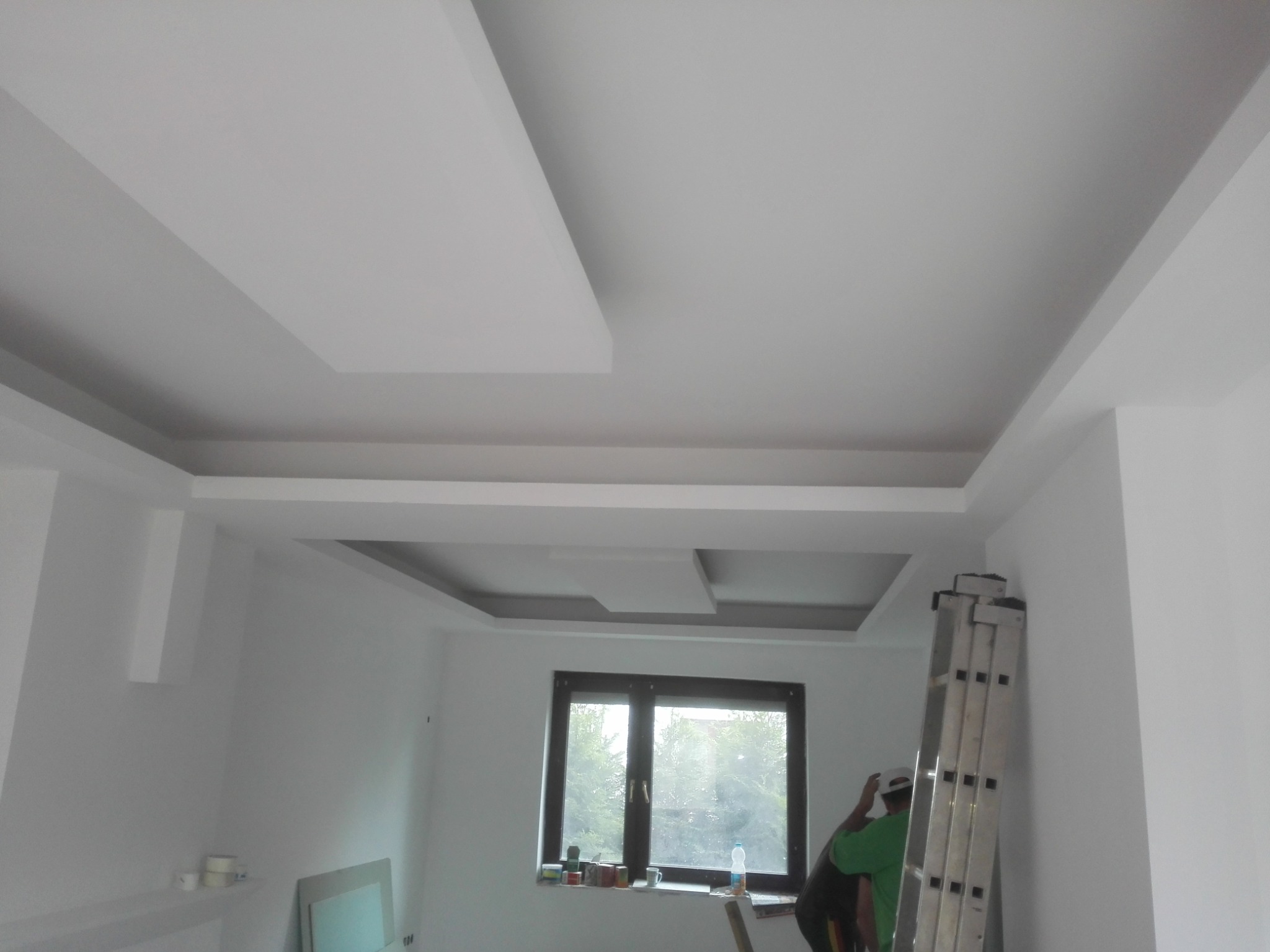 Nowoczesny sufit podwieszany z elementami dekoracyjnymi, widoczny fragment pomieszczenia w trakcie prac wykończeniowych, drabina oparta o ścianę, pracownik w tle.