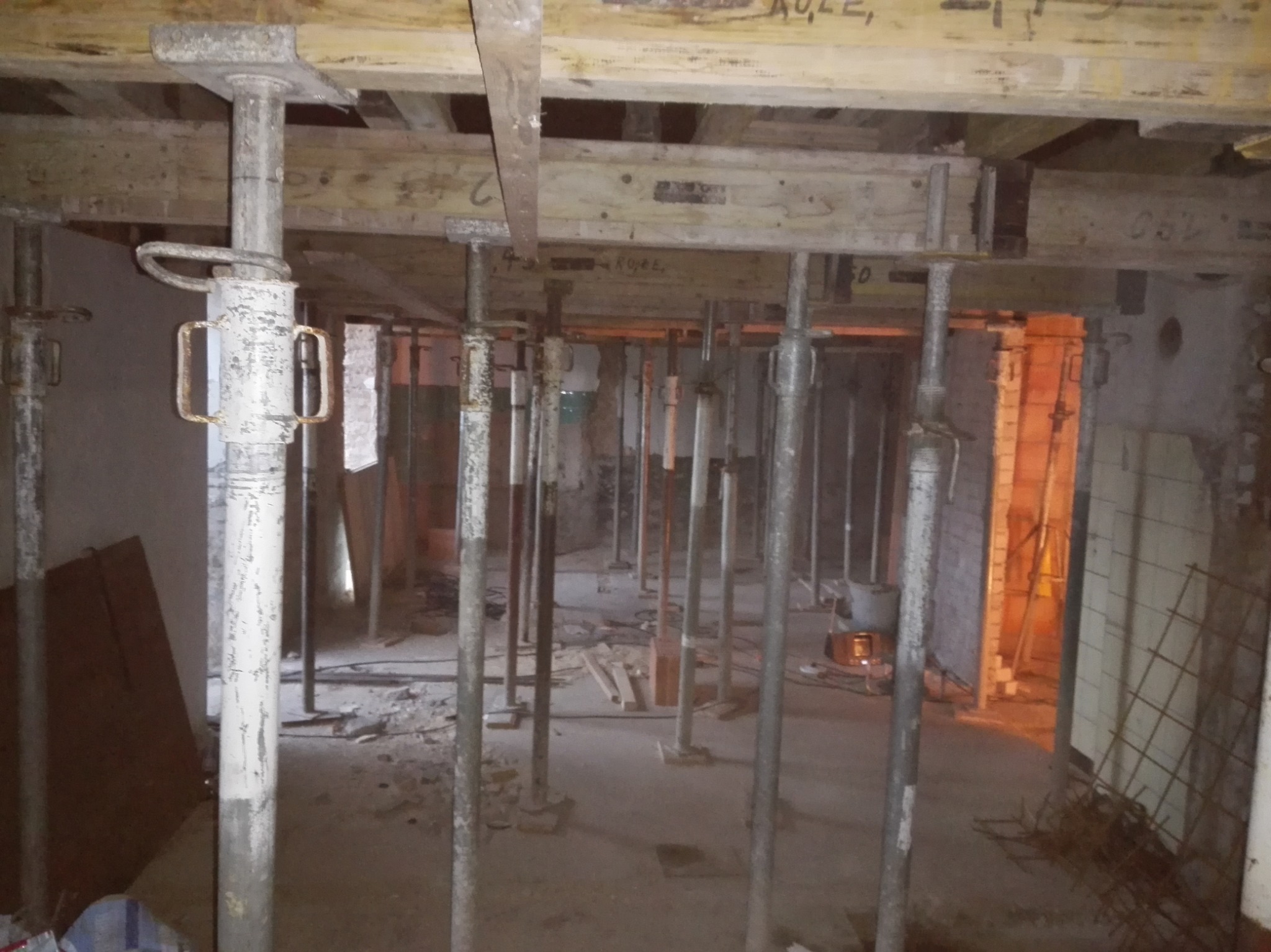 Wnętrze w trakcie budowy z widocznymi podporami stropowymi, drewnianymi belkami szalunkowymi i porozrzucanymi materiałami budowlanymi. Widoczne ślady po tynku na ścianach.
