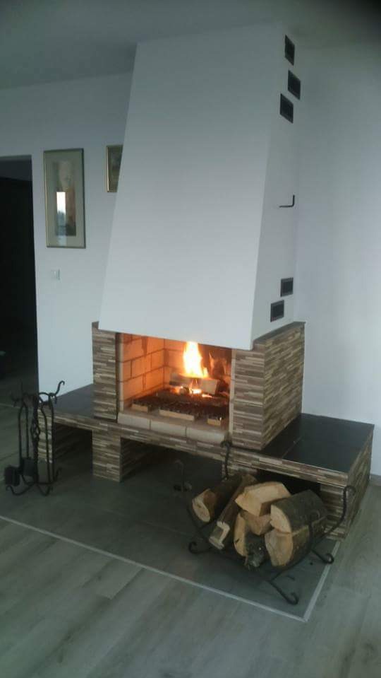 Nowoczesny kominek z wkładem obudowanym płytkami imitującymi kamień, z paleniskiem wypełnionym płonącym drewnem, na podłodze stojak z drewnem, a na ścianie obrazy w złotych ramach.