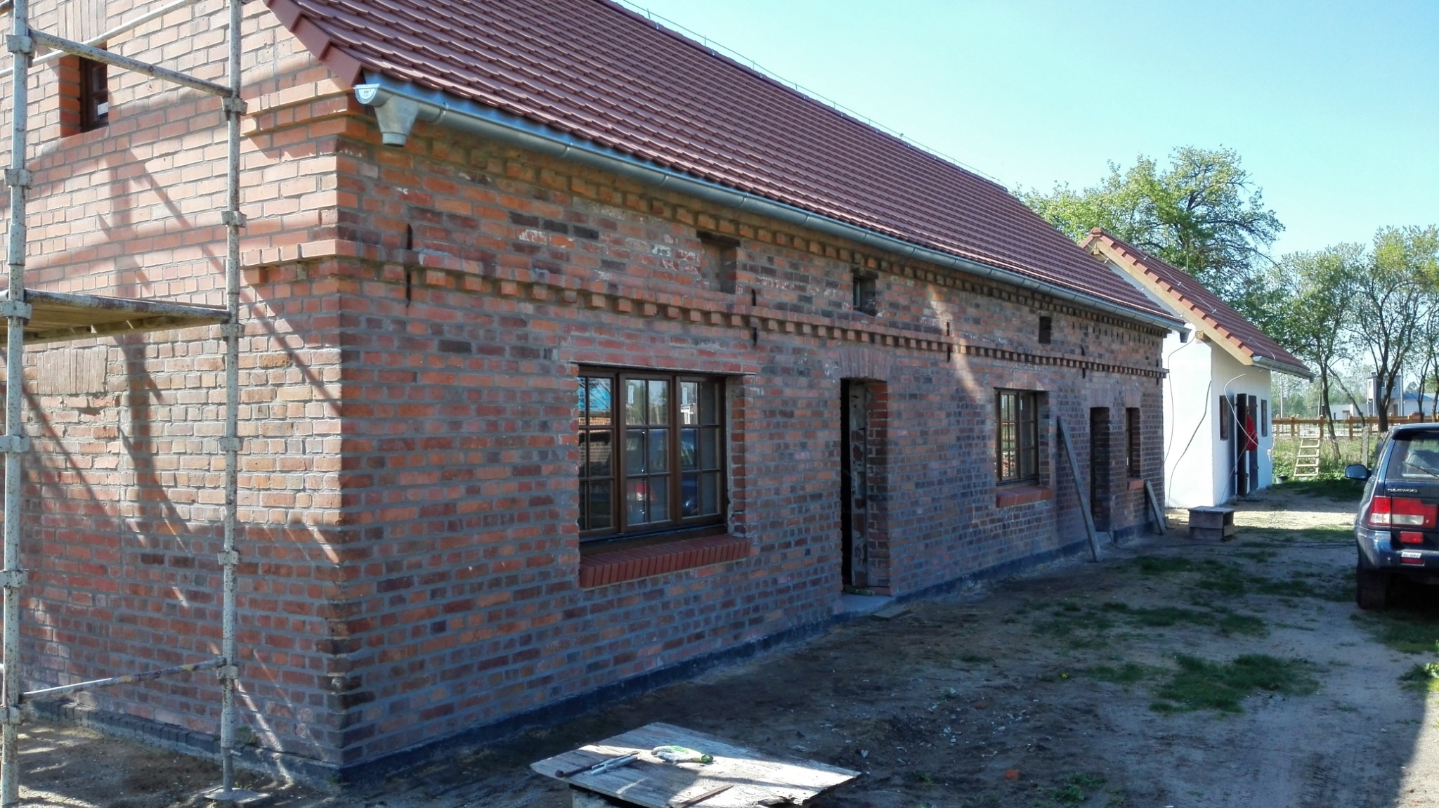 Surowa, czerwona elewacja z cegły klinkierowej nowo wybudowanego domu z widocznymi oknami i drzwiami, częściowo zasłonięta rusztowaniem, z fragmentem dachu z czerwonej dachówki i widoczną rynną.