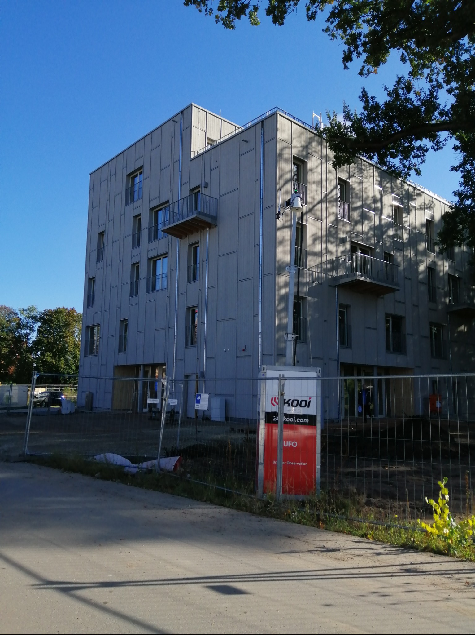 Wielopiętrowy budynek mieszkalny z szarej elewacji panelowej, z małymi balkonami, widoczny zza ogrodzenia budowy i kontenera z logo firmy.