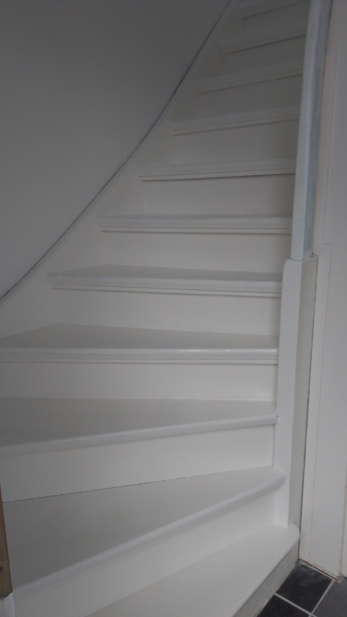 Białe, drewniane schody prowadzące na górę, z widoczną ścianą i fragmentem podłogi z ciemnych płytek.