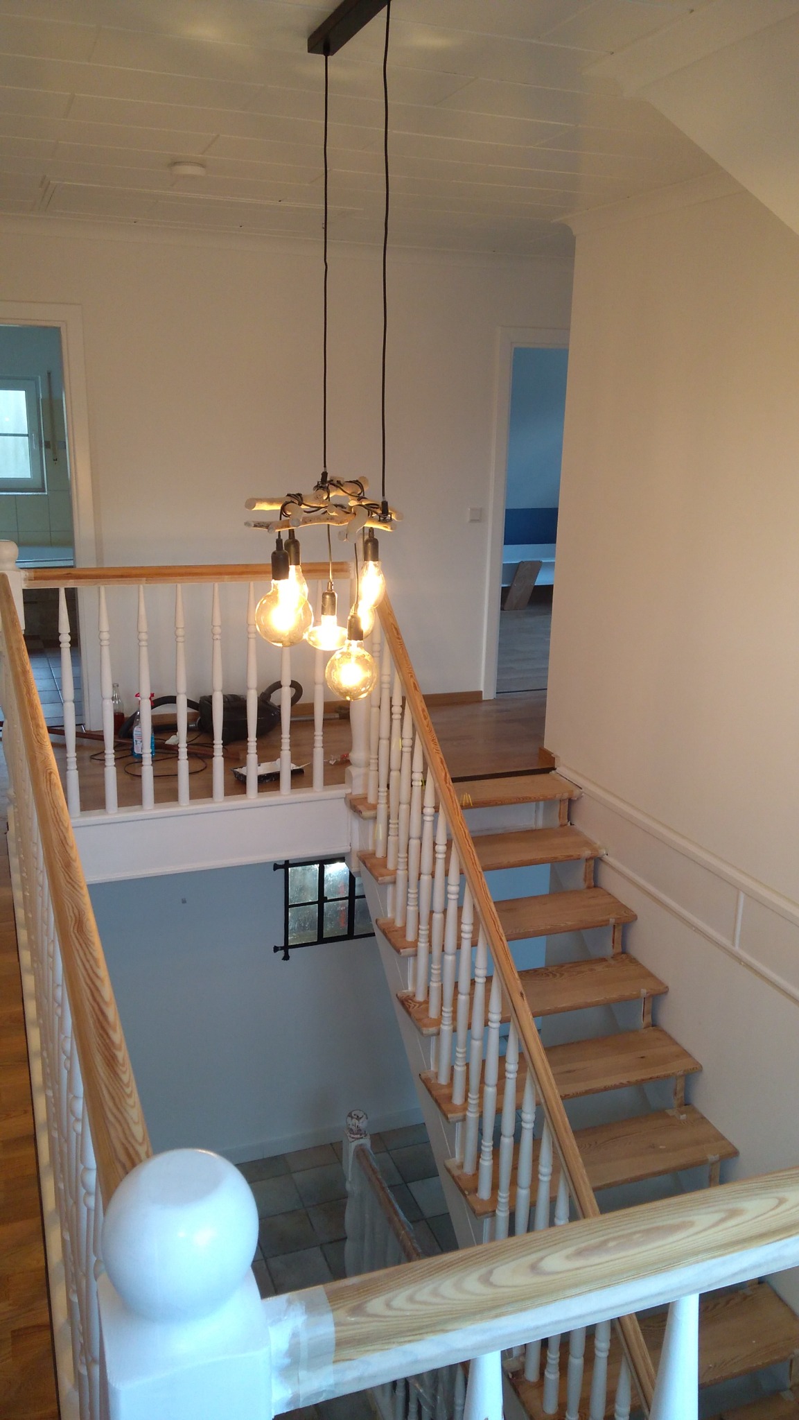 Wnętrze domu w trakcie wykańczania: drewniane schody z białą balustradą, designerska lampa wisząca z żarówkami Edisona, widok z góry na antresolę z widocznymi narzędziami pracy.