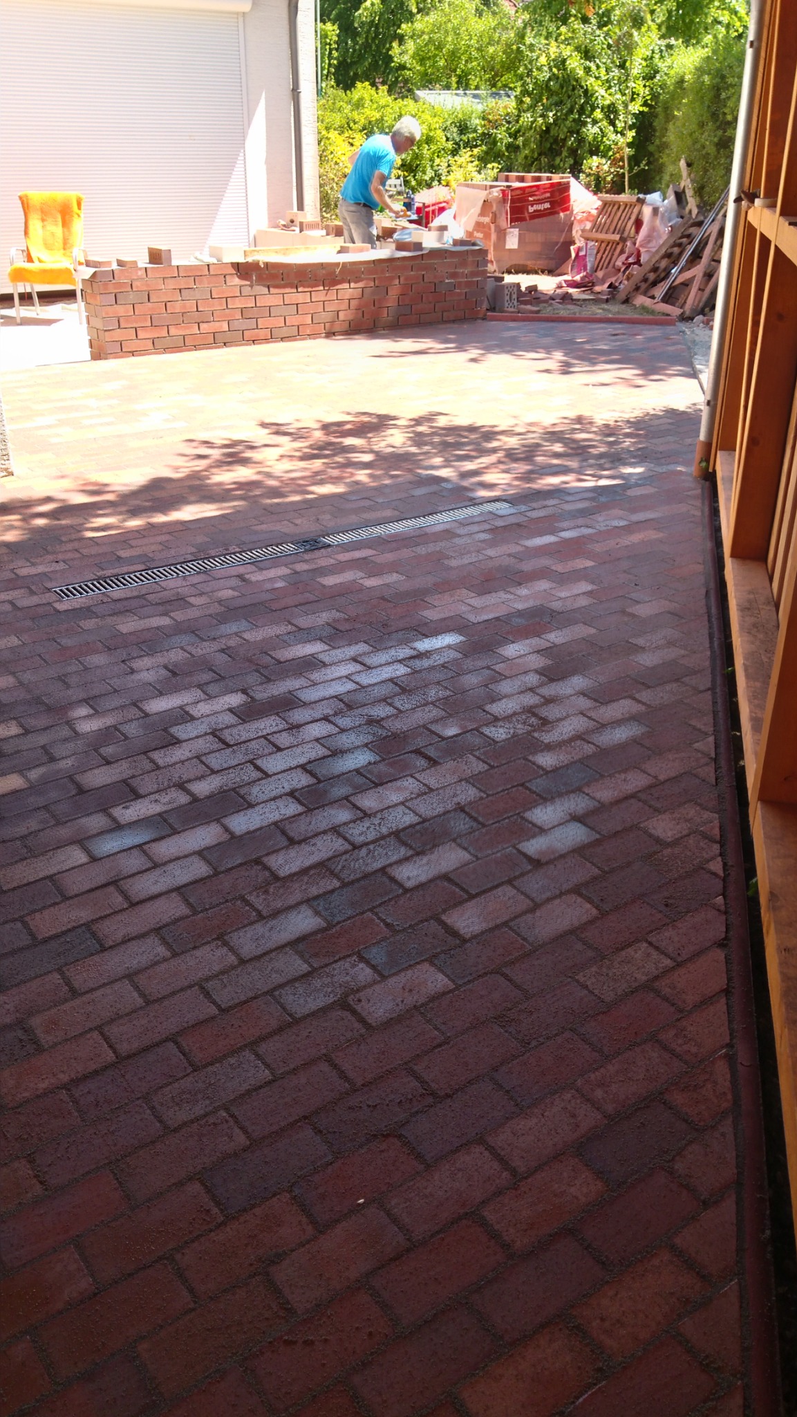 Murarz w niebieskiej koszulce w trakcie wznoszenia niskiego muru z czerwonej cegły na zewnątrz budynku, widok z góry na brukowaną powierzchnię.