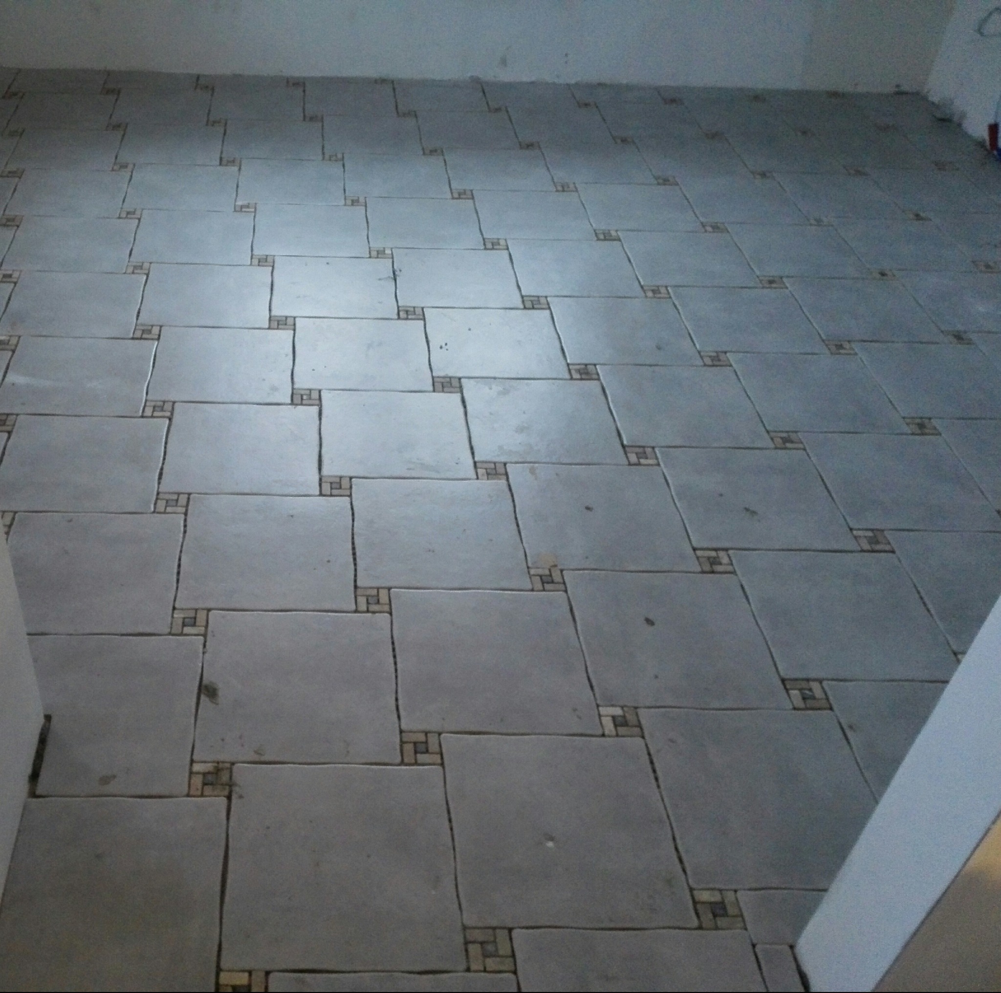 Ułożona podłoga z szarych, kwadratowych płytek z dekoracyjnymi, mozaikowymi wstawkami w rogach, widok z góry.