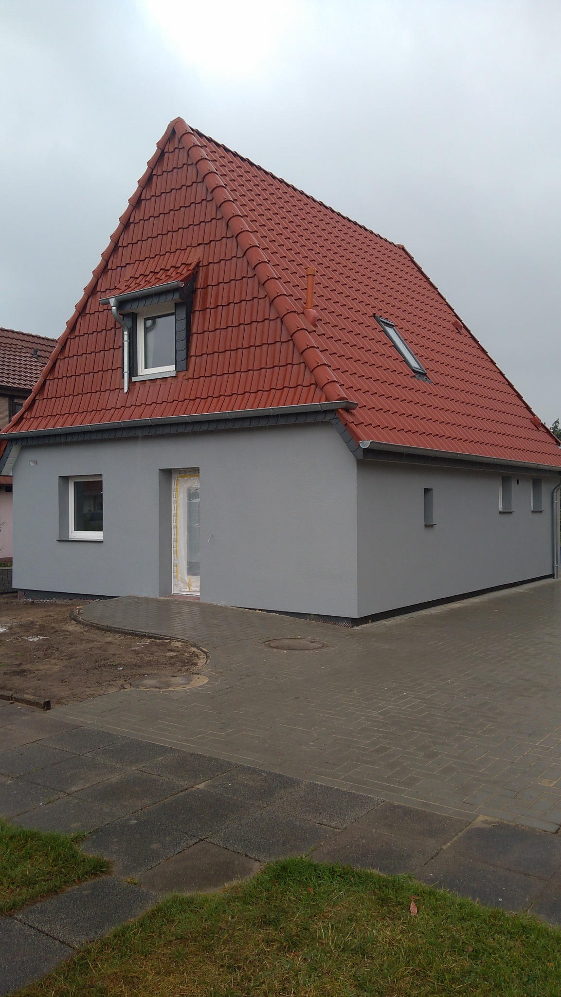 Nowoczesny dom z szarą elewacją i czerwoną dachówką, widoczne okna dachowe i rynny, teren wokół domu częściowo wyłożony kostką brukową, a częściowo przygotowany pod trawnik.