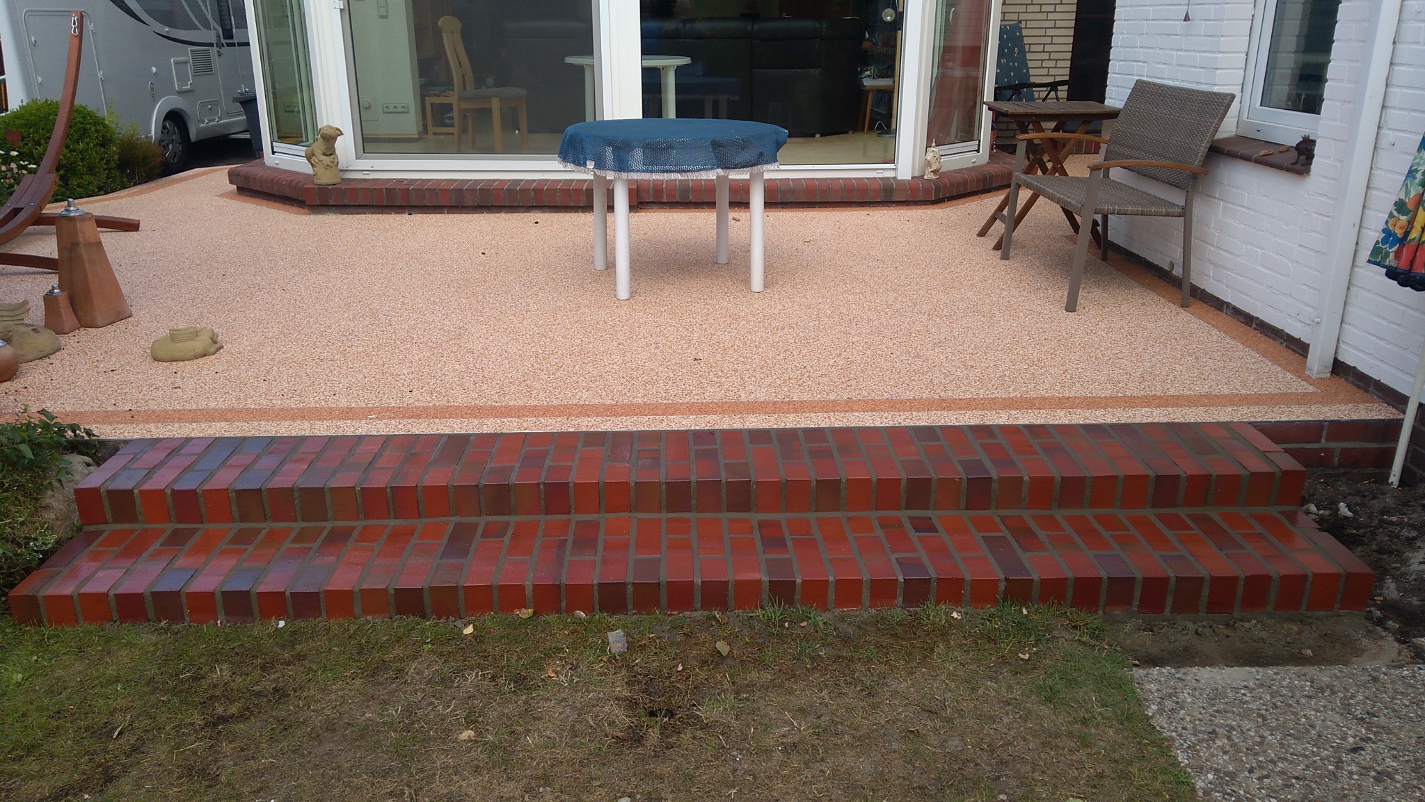 Wykończony taras z żywicy epoksydowej w kolorze brzoskwini, z dwustopniowymi schodami z czerwonej cegły, białymi drzwiami tarasowymi i meblami ogrodowymi.