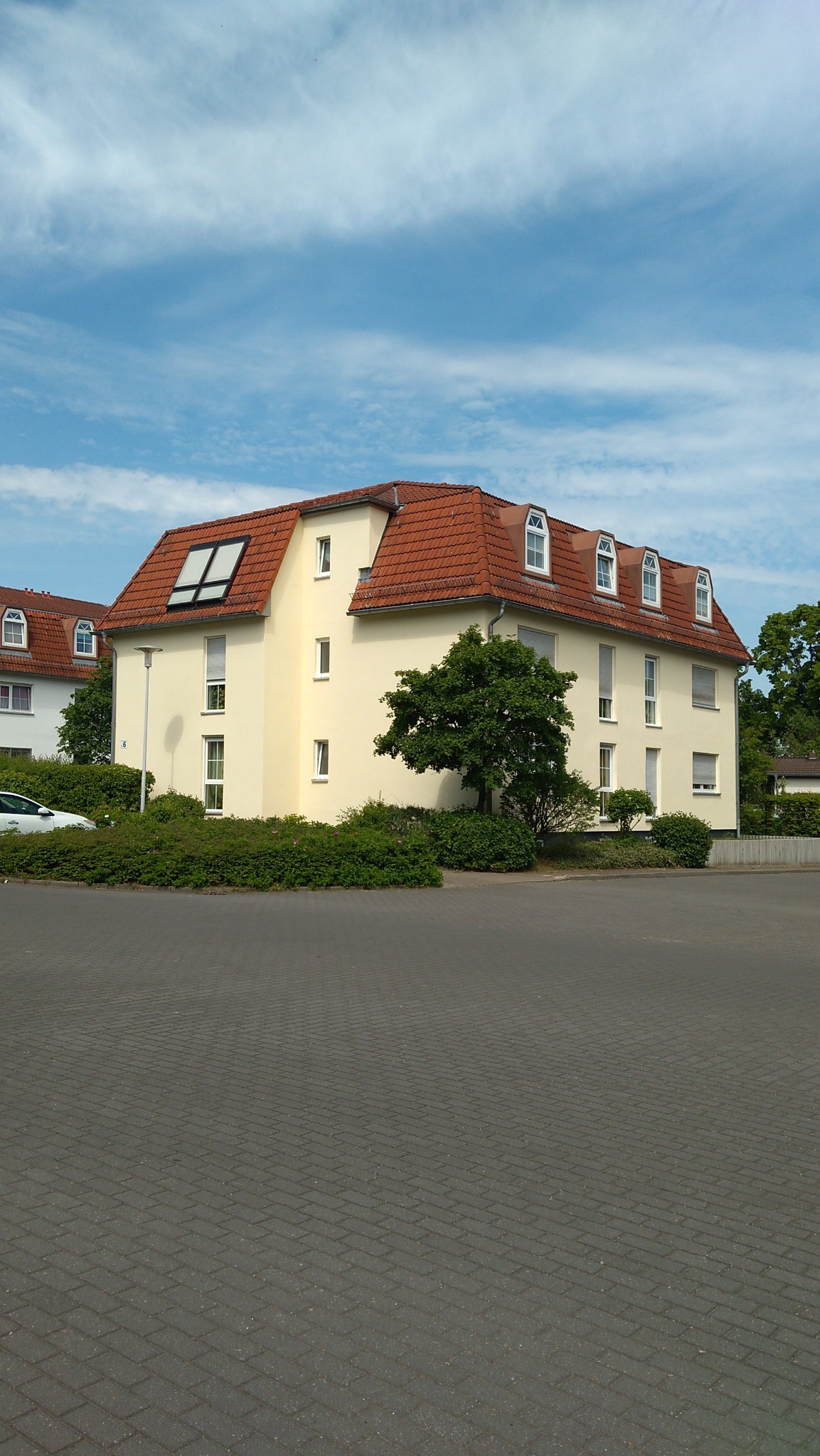 Żółty budynek mieszkalny z czerwonym dachem, widoczne okna dachowe i lukarny, otoczony zielenią, na pierwszym planie brukowany plac.