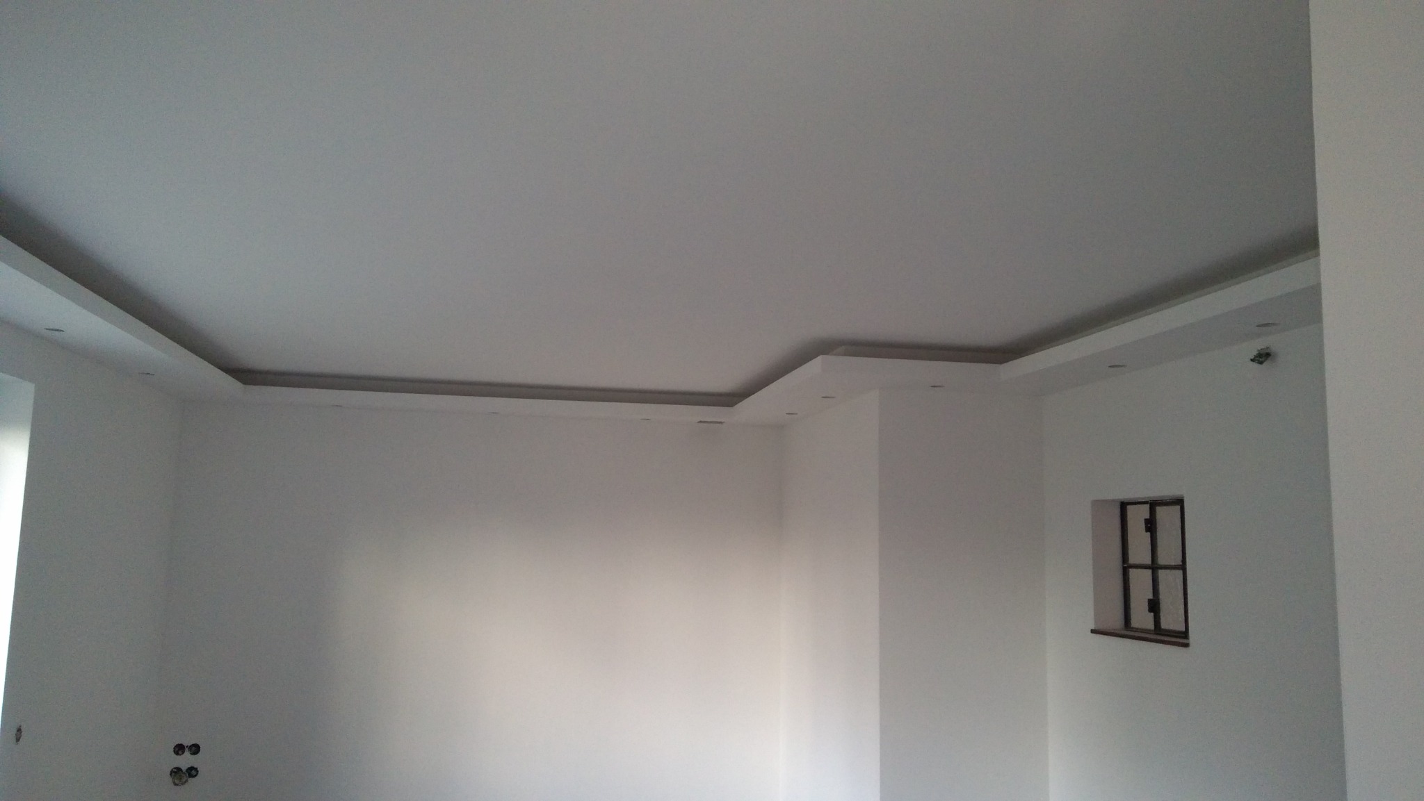 Białe wnętrze z zamontowaną, podwieszaną antresolą z oświetleniem punktowym, widoczne okno z ciemną ramą.