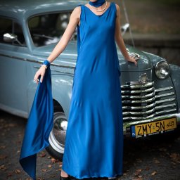 MARCIN FLAJSZER HANDEL I USŁUGI - Kobieta w długiej, kobaltowej sukni w stylu lat 30. pozuje przy zabytkowym, jasnoniebieskim samochodzie z chromowanymi elementami, trzymając w ręku pasujący szal.