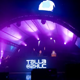 MARCIN FLAJSZER HANDEL I USŁUGI - DJ Talla 2XLC na scenie w Gorzowie Wielkopolskim, oświetlonej fioletowymi światłami, z fotografem u dołu kadru. Widoczna konstrukcja sceny.