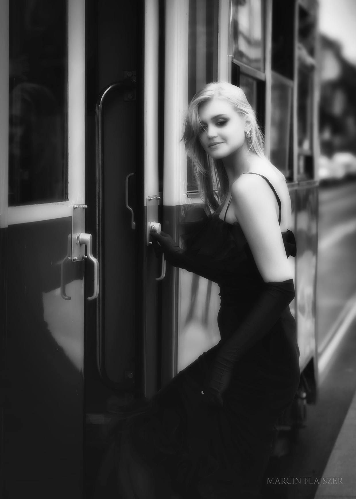 Czarno-białe zdjęcie eleganckiej kobiety w długiej sukni i rękawiczkach, otwierającej drzwi wagonu tramwajowego w stylu retro.