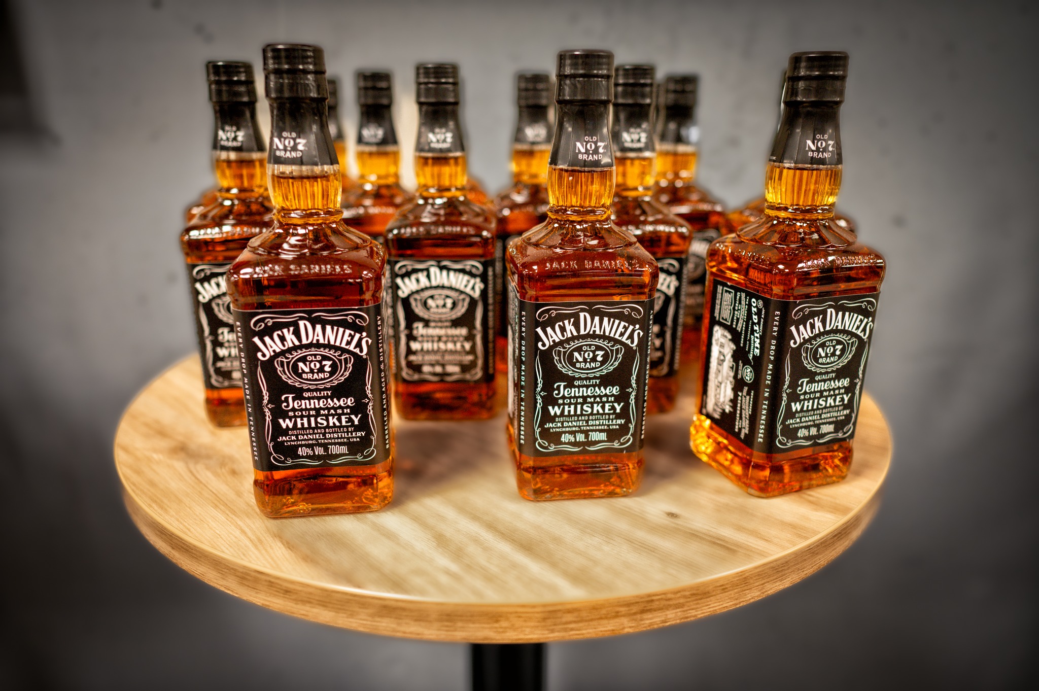 Precyzyjnie oświetlony, symetryczny układ kilkunastu butelek Jack Daniel's na okrągłym, drewnianym stoliku z czarną nogą, tworzący efektowną ekspozycję. Głębokie cienie podkreślają kształt butelek.