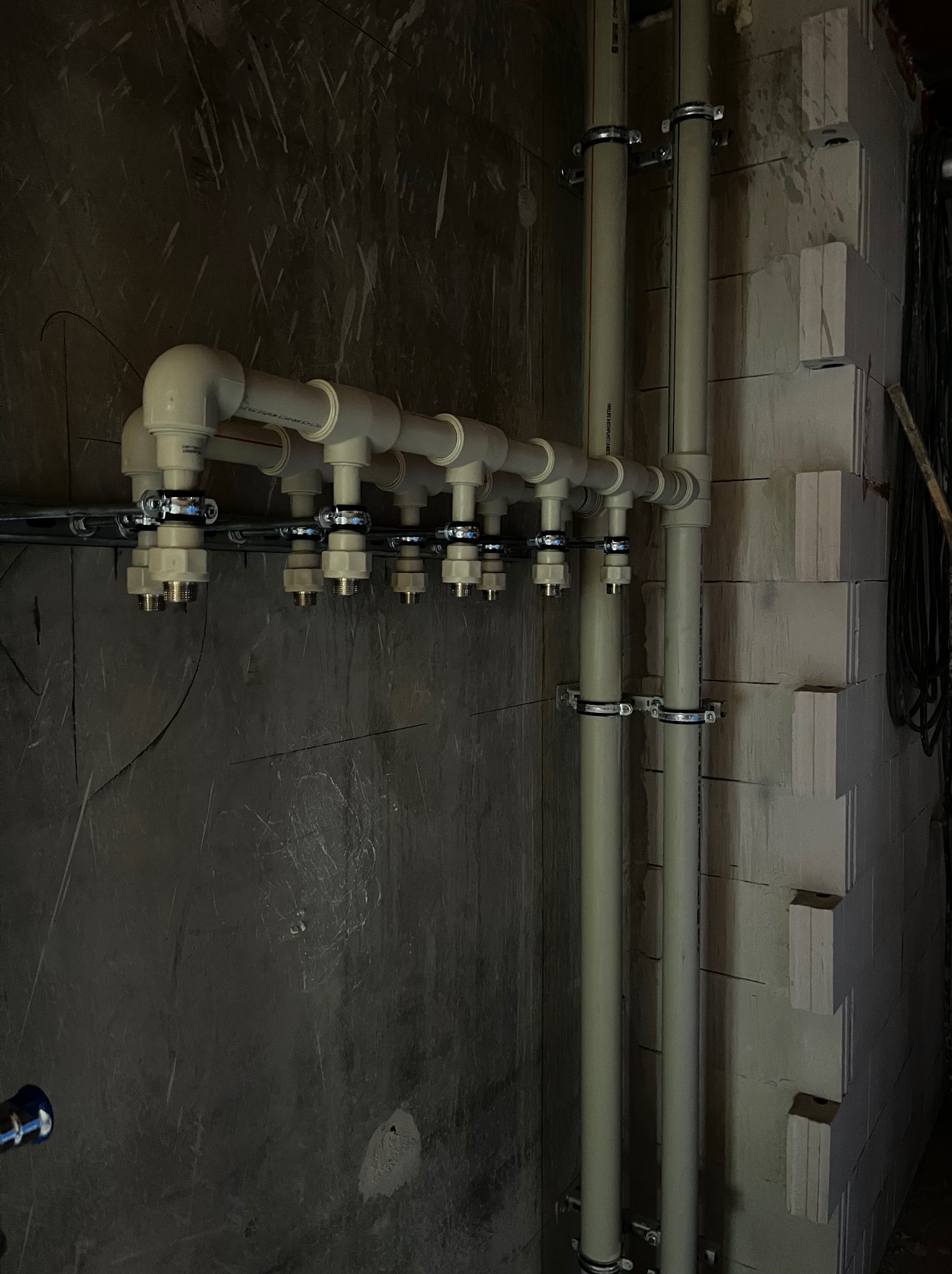 Rozdzielacz instalacji hydraulicznej z rurami PCV zamocowany do ściany z bloczków betonowych, widoczne metalowe złączki i obejmy.