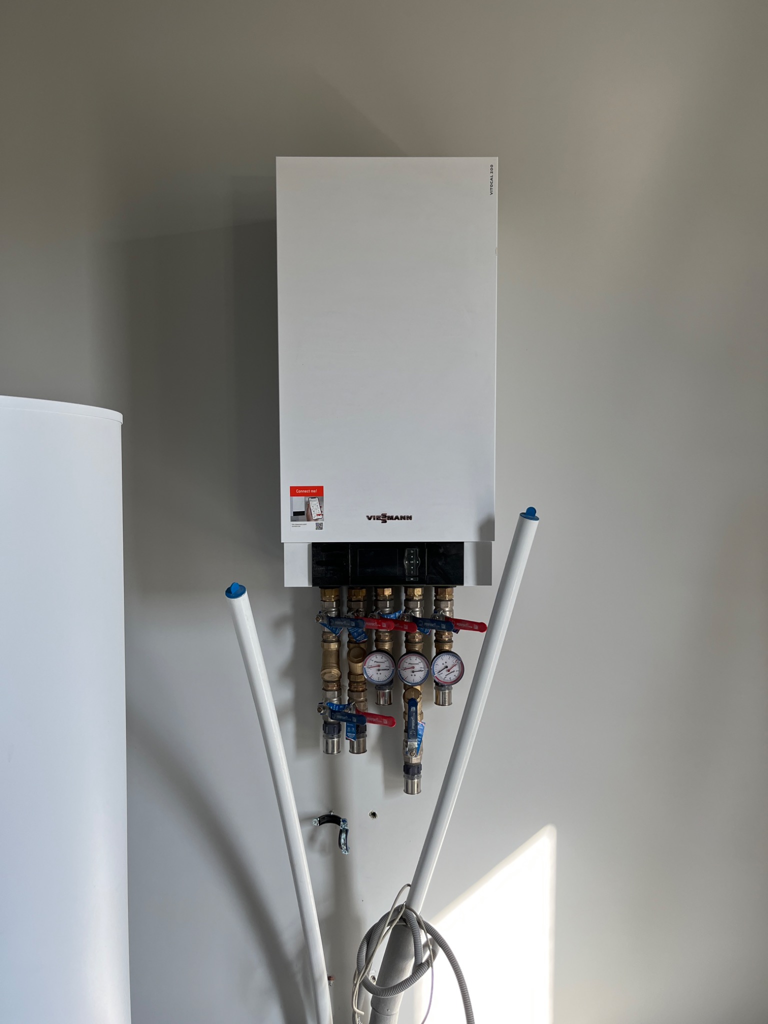 Biały, wiszący kocioł grzewczy Viessmann z podłączonymi rurami i manometrami na szarej ścianie, widoczny również biały zasobnik wody.