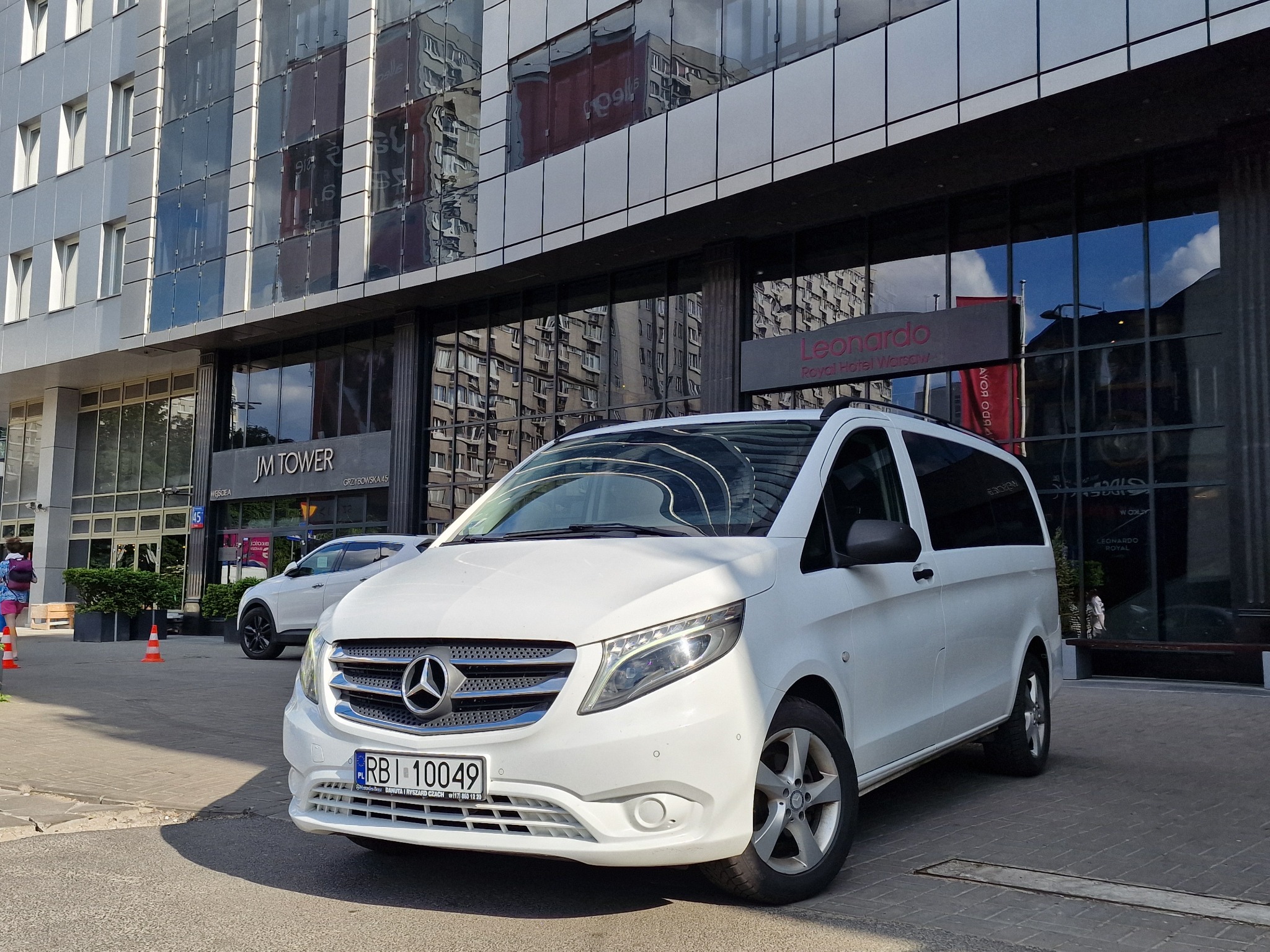 Biały Mercedes Vito zaparkowany przed wejściem do hotelu Leonardo Royal w Warszawie, z widoczną tablicą rejestracyjną i odbiciem budynku w szybach.