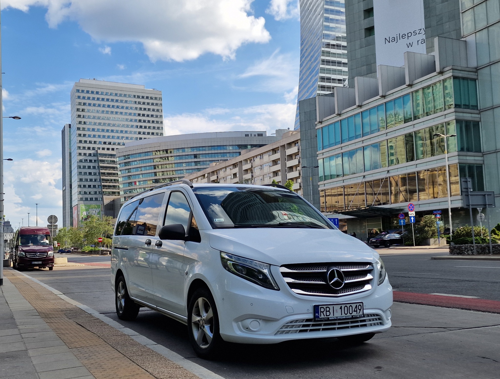 Biały Mercedes Vito zaparkowany na ulicy w centrum miasta, z widocznymi wieżowcami i budynkami biurowymi w tle.