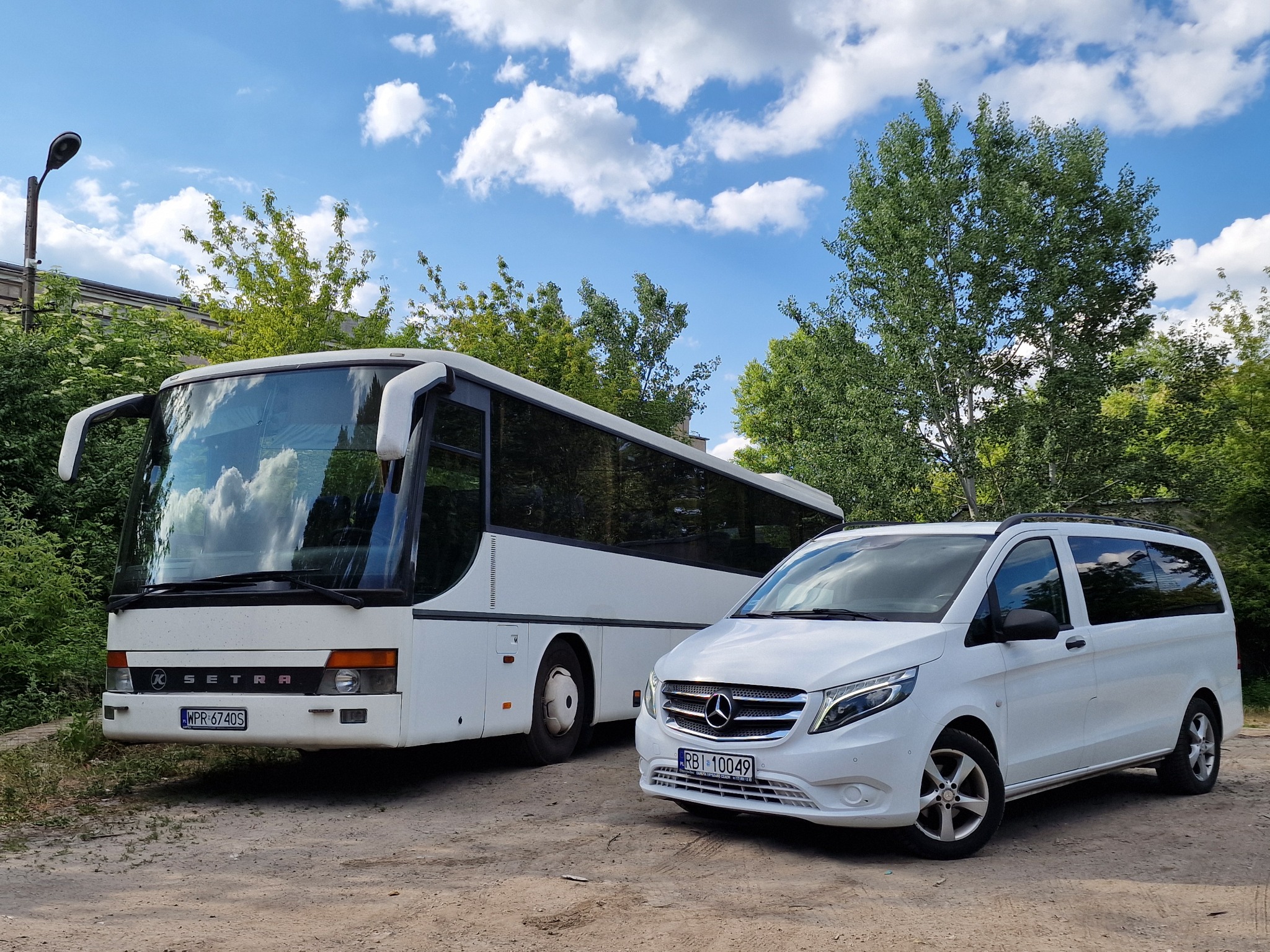 Dwa białe pojazdy: autokar Setra z przodu i minibus Mercedes Vito z tyłu, zaparkowane na żwirowym placu w otoczeniu zieleni, przy słonecznej pogodzie z chmurami na niebie.
