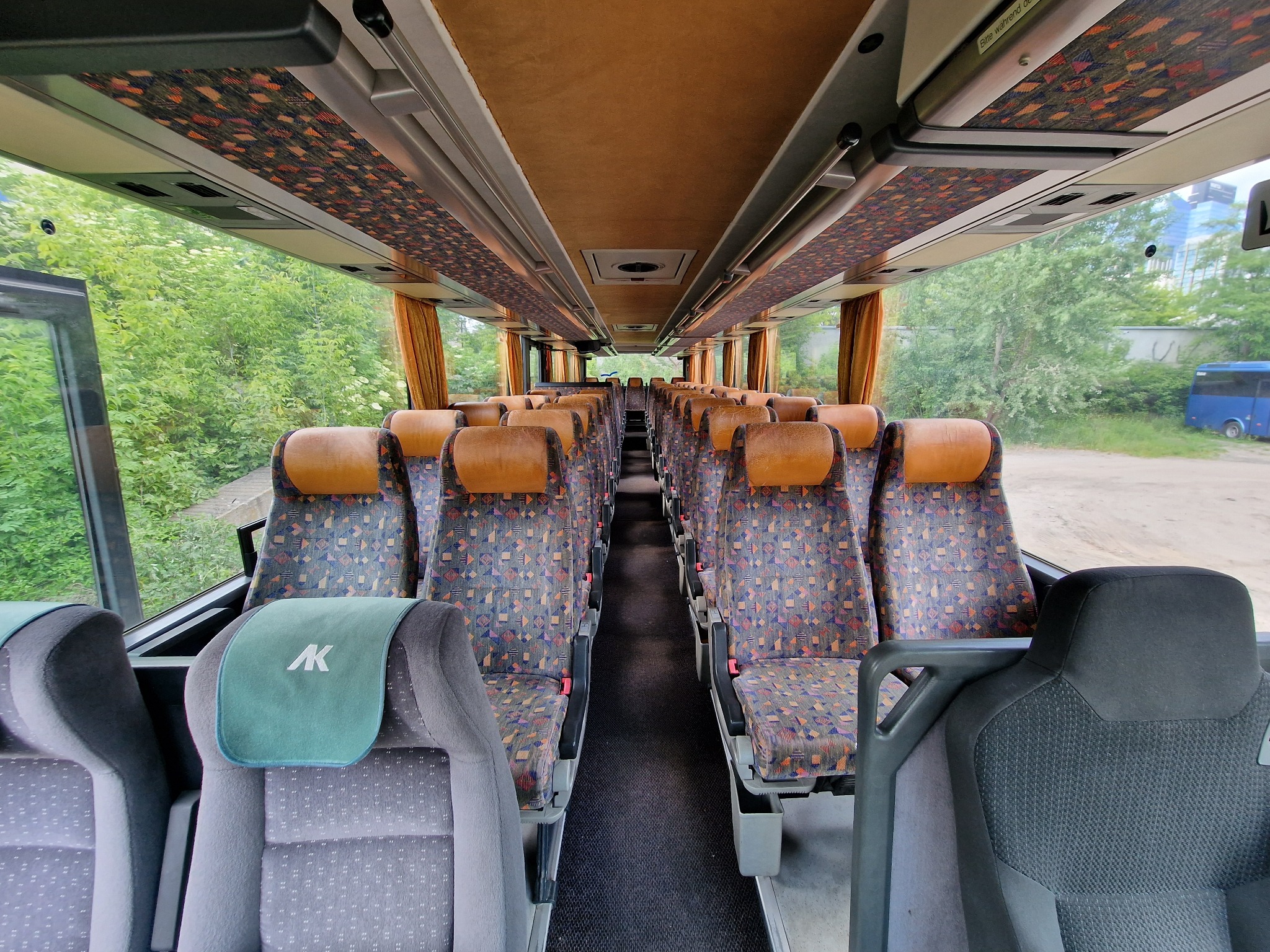 Wnętrze autobusu z rzędami foteli z brązowymi zagłówkami i wzorzystą tapicerką, widok z przodu pojazdu, przez otwarte drzwi widać zieleń drzew.