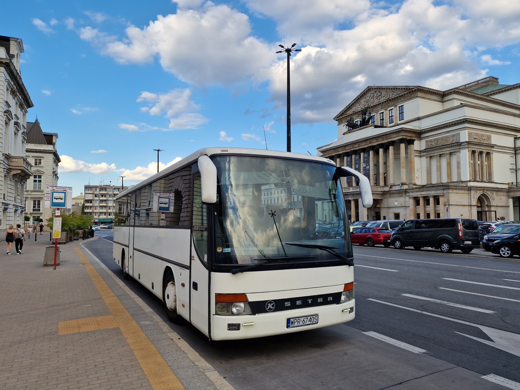 Biały autokar marki Setra zaparkowany na ulicy z widocznym Teatrem Wielkim w Warszawie w tle, odbicie budynku w szybie autobusu.