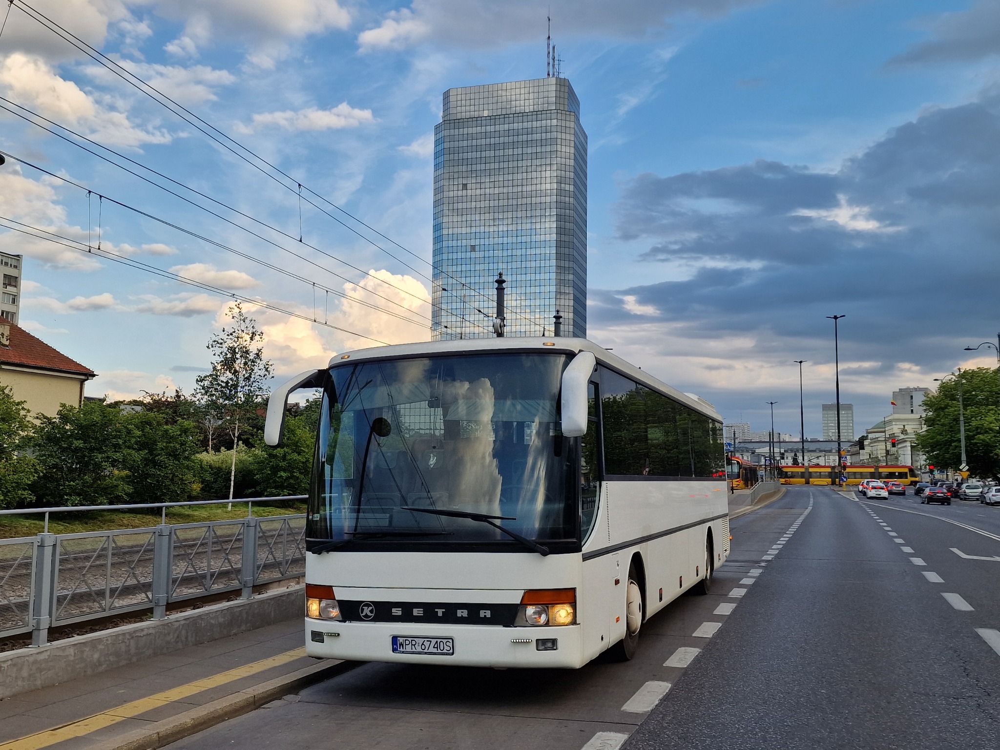 Biały autokar Setra na tle miejskiego krajobrazu z nowoczesnym wieżowcem odbijającym chmury, widoczne linie tramwajowe i sygnalizacja świetlna.
