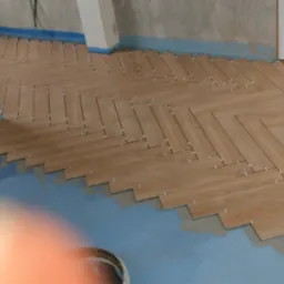 Układanie parkietu w jodełkę z płytek ceramicznych imitujących drewno, widoczne krzyżyki dystansowe i fragment przygotowanego podłoża w pomieszczeniu w stanie surowym.