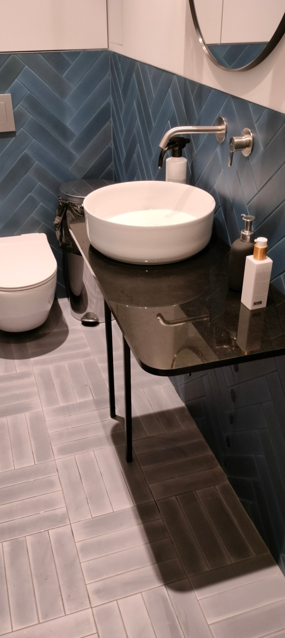 Nowoczesna łazienka z umywalką nablatową na czarnym blacie, armaturą w kolorze stali, niebieskimi płytkami ułożonymi w jodełkę na ścianie i szarymi płytkami na podłodze, z widoczną toaletą i koszem...