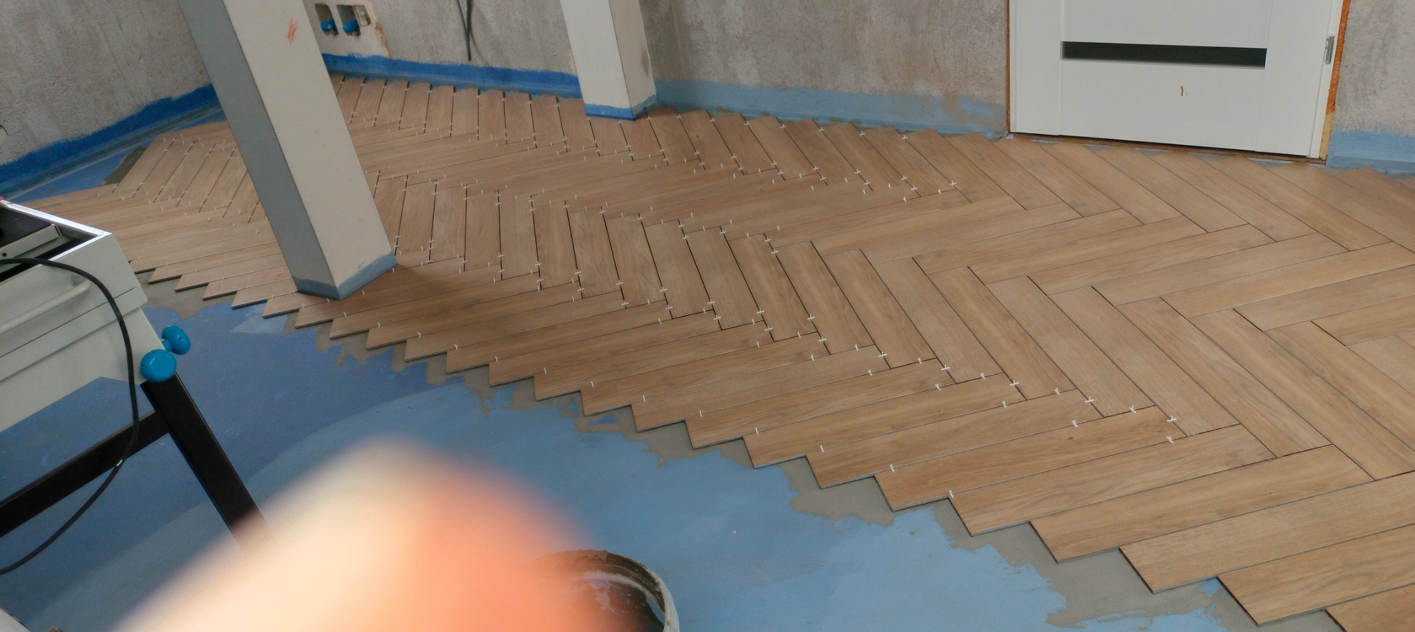 Układanie parkietu w jodełkę z płytek ceramicznych imitujących drewno, widoczne krzyżyki dystansowe i fragment przygotowanego podłoża w pomieszczeniu w stanie surowym.