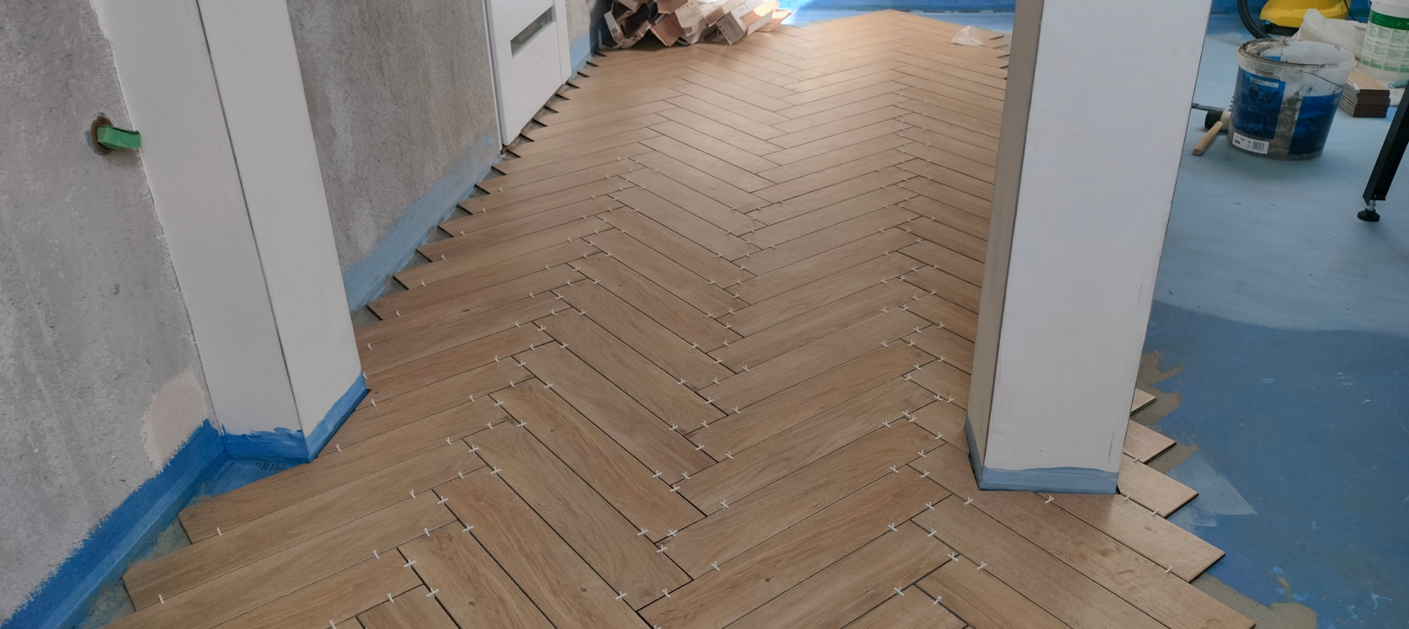 Układanie płytek podłogowych w jodełkę, widoczne krzyżyki dystansowe, zabezpieczone folią ściany i podłoga, w tle wiadro z klejem i zapas płytek.