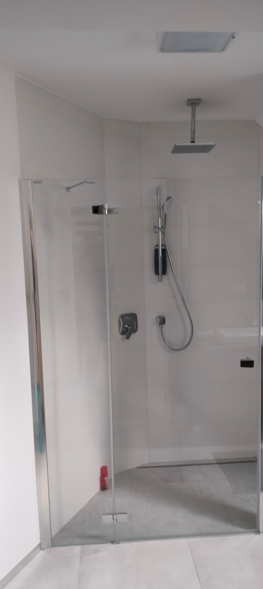 Nowoczesna kabina prysznicowa z deszczownicą sufitową i szarymi płytkami, widoczna butelka czerwonego płynu do czyszczenia w rogu.