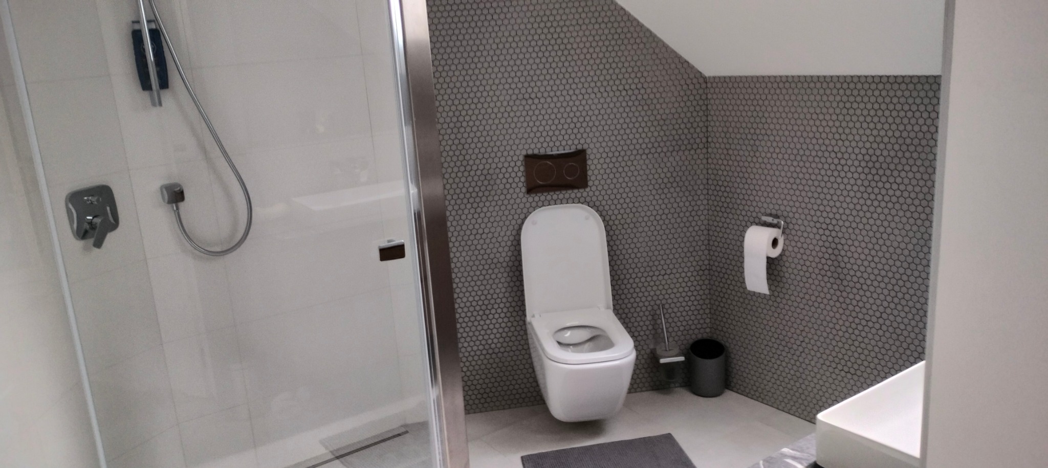 Nowoczesna łazienka z kabiną prysznicową ze szklanymi drzwiami, toaletą wiszącą na ścianie z szarą mozaiką i akcesoriami w odcieniach szarości.